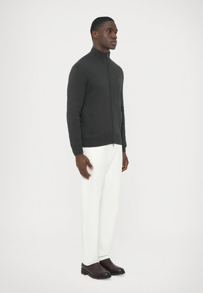 Sötétszürke bordázott pulóver magas gallérral, cipzáras záródással és oldalzsebekkel, fehér nadrággal és fekete bebújós cipővel párosítva.