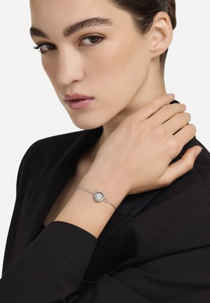 Hõbedast käevõru, millel on ümmargune ripats tekstuuriga pinnaga, kantud mudeli randmel musta bleiseriga kombineeritult.