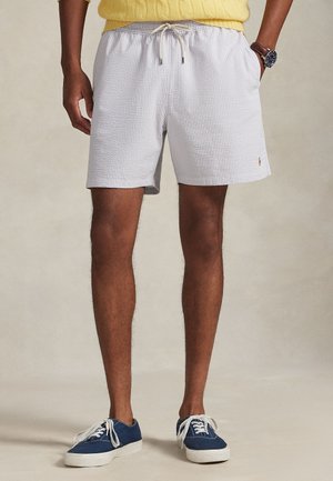 Mand iført hvide, teksturerede shorts med snøre, gul trøje, blå sneakers og armbåndsur, stående med hænderne i lommerne.