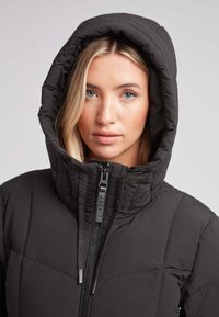 Chaqueta negra acolchada con un alto cuello y una capucha. Presenta un cierre delantero y una textura acolchada, enfatizando la calidez y el aislamiento.