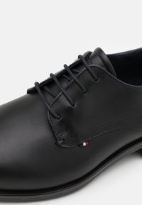 Chaussure habillée en cuir noir avec surface lisse, bout rond, design à cinq œillets et lacets, et coutures ton sur ton. Présente un petit logo en contraste de couleur.