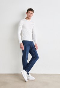 Hvit langermet skjorte, blå jeans og hvite joggesko. Skjorten har en liten logo, og jeansene har en klassisk passform med diskre søm.