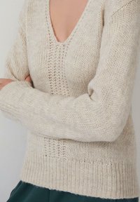 Persona che indossa un maglione lavorato a maglia beige a scollo a V con le braccia incrociate e pantaloni verde scuro su uno sfondo neutro.