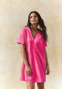 Īsa rozā kleita no viegla auduma. Tai ir V veida kakla izgriezums un īsas piedurknes ar smalku tekstūru un brīvu piegriezumu.