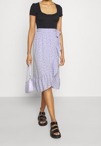 Musta bodysuit neliön muotoisella kaula-aukolla yhdistettynä laventelin väriseen, kukkakuosisella, frillareunallisella kietaisuhameella. Malli pitää kädessään pientä laventelin väristä laukkua ja on pukeutunut mustiin sandaaleihin.