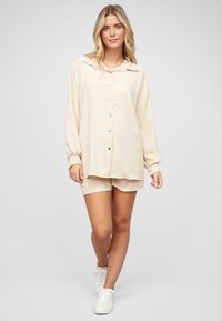 Beige oversized skjorte med knaplukning, lange ærmer med elastiske manchetter, parret med matchende shorts. Modellen har hvide sneakers på.