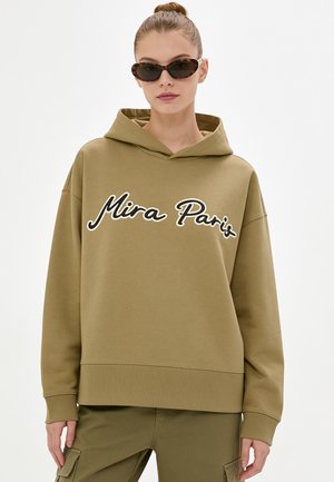 Donna che indossa una felpa con cappuccio verde oliva con il testo "Mira Paris", pantaloni cargo abbinati e occhiali da sole cat-eye tartarugati, capelli raccolti in uno chignon.
