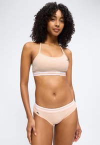 Γυναίκα με σγουρά μαλλιά, φορώντας μπεζ αθλητικό σουτιέν και αντίστοιχο σλιπ με λευκές ριγέ ελαστικές ταινίες, στέκεται μπροστά σε απλό φόντο.