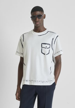 Joven con gafas de sol negras, camiseta blanca con detalles de bolsillo y costuras en estilo boceto negro, y pantalones azul oscuro, de pie con una mano en el bolsillo.