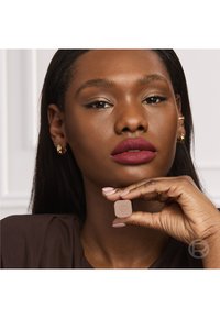 Frau mit dunkler Haut und rotem Lippenstift hält ein kleines beiges Kosmetikprodukt nahe ihrem Kinn, trägt goldene Ohrringe und ein braunes Oberteil.