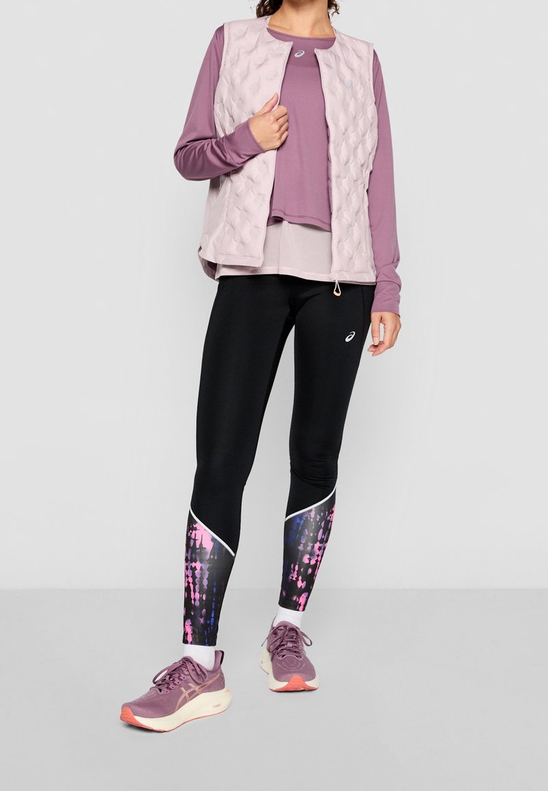 Gilet leggero trapuntato di un rosa pallido sopra una maglietta a maniche lunghe color malva. Leggings neri con un vivace motivo digitale rosa e blu, abbinati a sneakers viola.