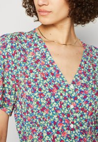 Femme aux cheveux bouclés portant une robe bleue à fleurs rouges, blanches et vertes, manches courtes bouffantes, un décolleté en V et un fin collier en or.