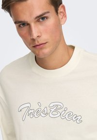 Kremowa bluza z miękkiej bawełny, z nadrukiem graficznym z napisem "Très Bien" w stylowej czcionce kursywą.