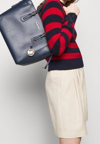 Granatowy skórzany plecak z złotym logo MK, wyposażony w dwa paski na ramiona i klasyczną konstrukcję. Model nosi czerwony i granatowy sweter w paski oraz beżowe szorty.