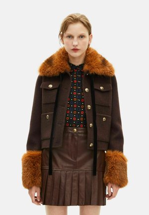 Brun uldjakke med en imiteret pelskrave og manchetter i orange, prydet med guldfarvede knapper og to forlommer, lagd over en mønstret bluse og nederdel.