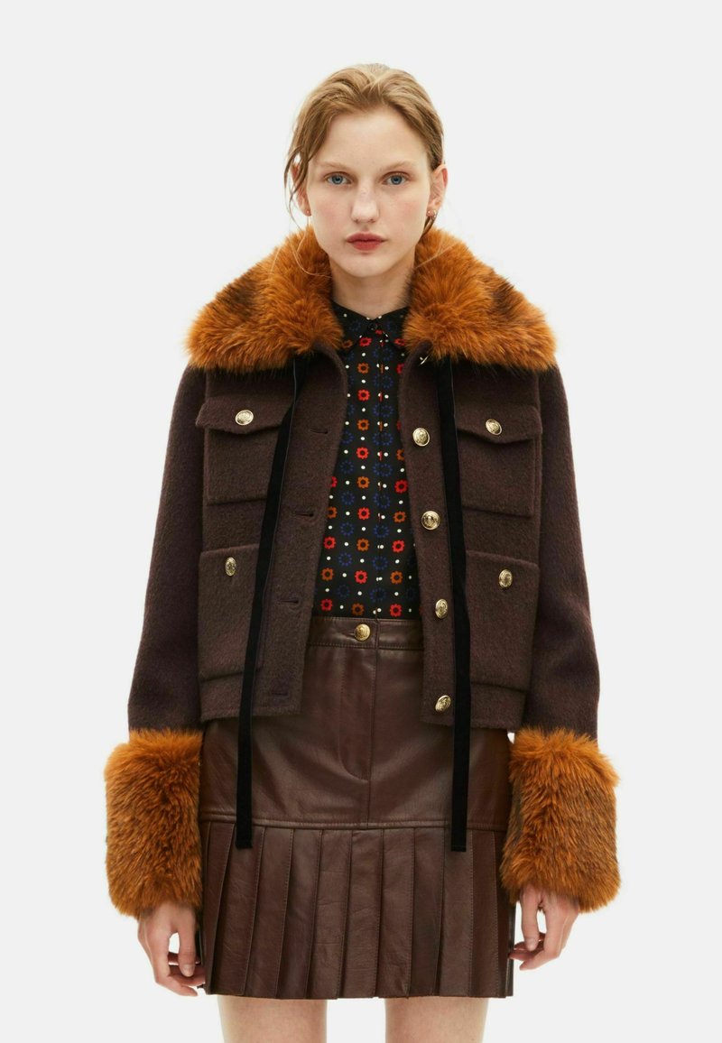 Chaqueta de lana marrón con un cuello y puños de piel sintética naranja, adornada con botones dorados y dos bolsillos frontales, superpuesta a una blusa y falda estampadas.