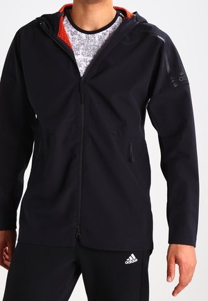 Mann trägt schwarzen Adidas Zip-Hoodie mit rotem Innenfutter und schwarze Adidas Trainingshose, steht vor einem einfachen Hintergrund.