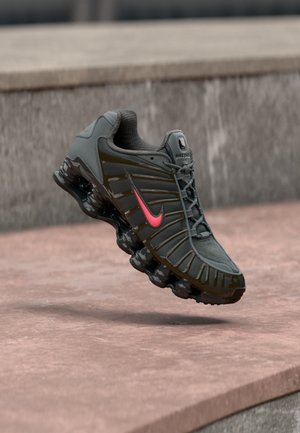 Ciemnoszary but do biegania Nike z czerwonym logo i segmentowaną amortyzowaną podeszwą zawieszony nad kamiennymi schodami.