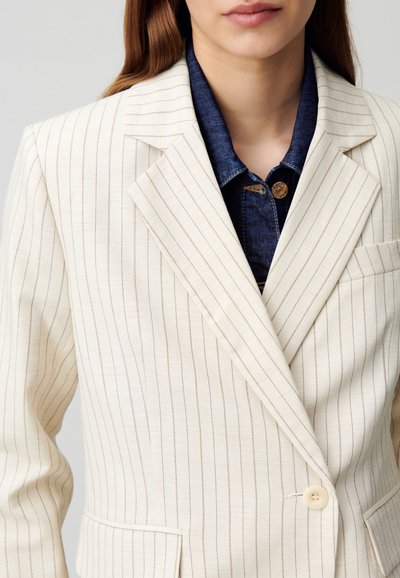Blazer beige a righe con un solo bottone, revers a scendere e taschino sinistro sul petto, indossato sopra una camicia di denim con dettaglio a bottone.