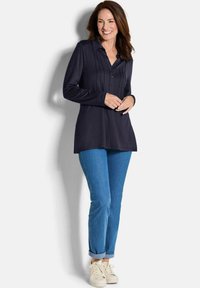 Camicia a maniche lunghe blu marina con patta abbottonata e dettagli a pieghe, abbinata a jeans azzurri arrotolati e sneakers bianche.