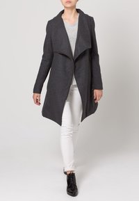 Grauer Wollmantel mit asymmetrischer Vorderseite, großen Revers und Reißverschluss, kombiniert mit einem hellen Pullover und weißen Hosen, getragen mit schwarzen Stiefeln.