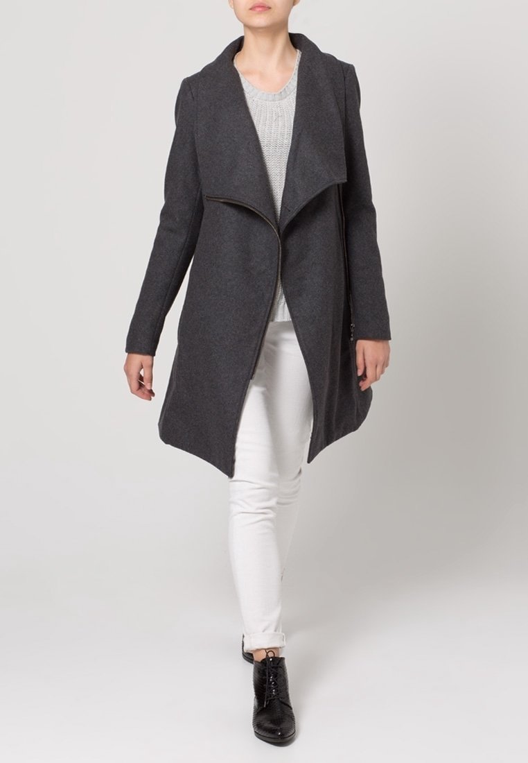 Grauer Wollmantel mit asymmetrischer Vorderseite, großen Revers und Reißverschluss, kombiniert mit einem hellen Pullover und weißen Hosen, getragen mit schwarzen Stiefeln.