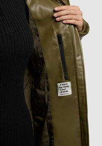 Olijfgroene leren jas met een ritszak aan de binnenkant en een stoffen label waarop staat: "Een warme omhelzing van stof."