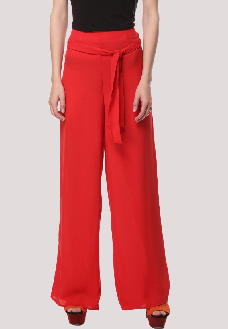 Mujer con pantalones rojos de pierna ancha y cintura alta, con lazo en la cintura, combinados con una blusa negra y sandalias de plataforma naranjas.