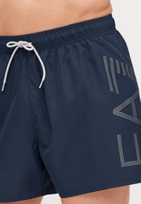 Shorts de bain bleu marine en tissu léger, dotés d'une taille élastique avec un cordon de serrage gris et des accents graphiques en strass blancs.