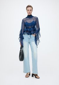 Γυναίκα που φοράει διάφανο μπλε σκούρο πουκάμισο με βολάν, ανοιχτό μπλε παντελόνι με φαρδιά μπάτζα, μαύρα πέδιλα με τακούνι, κρατώντας μαύρη τσάντα στο χέρι, πάνω σε λευκό φόντο.