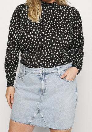 Kvinde iført en sort langærmet top med hvide uregelmæssige prikker og en lyseblå denimnederdel med frynset kant, hånd stukket i lommen.