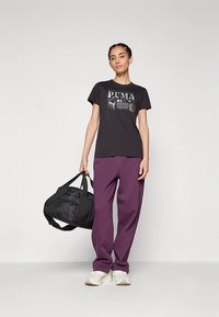 Černé tričko s grafickým logem, ve spojení s fialovými širokými kalhotami, držící černou sportovní tašku a obuté do bílých tenisek.