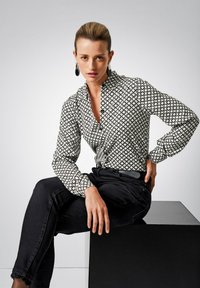 Black and white patterned shirt with a geometric design, featuring a collared neckline and long sleeves, worn with black jeans and a belt.