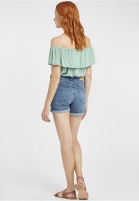 Minta zaļa off-the-shoulder blūze ar rāvējslēdzēja detaļām, saskaņota ar gaišziliem džinsa šortiem ar rullētiem malām. Modelis valkā tan krāsas sandales.