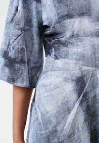 Indumento in denim con un motivo strutturato grigio e blu, design scomposto, maniche corte e dettagli di cucitura visibili.