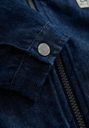 Detalj teksas jakne, tamnoplavi materijal, vidljivi šavovi, metalni zatvarač na klik, i centrirani zatvarač u kontrastnoj crnoj boji, s etiketom iznad.