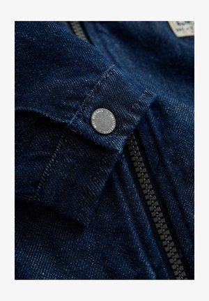 Teksariidest jakk detail, tumesinine kangas, nähtav õmblus, metallist trukk, ja keskel kontrastset musta värvi tõmblukk, ülal silt.