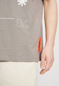 Harmaa T-paita kevyestä kankaasta, jossa on valkoisia graafisia kuvastoja, mukaan lukien numeroita ja oranssilla korostetut etiketit helman kohdalla.