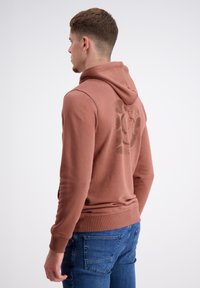 Bruine hoodie met een grote grafische print op de achterkant, geribbelde manchetten en zoom. Draagt met blauwe jeans. Zacht materiaal en casual ontwerp.