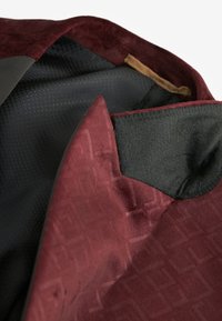 Gros plan sur le col d'une veste bordeaux avec une doublure noire texturée et des coussinets en tissu doux à l'intérieur de la zone du col.
