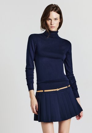 Femme portant un pull col roulé bleu marine et une jupe plissée avec une ceinture beige, se tenant contre un fond gris clair.