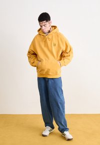 Sudadera con capucha de color amarillo mostaza, con un bolsillo frontal, ajuste de cordón y parche de logo, combinada con jeans de corte holgado en color azul y zapatillas blancas.