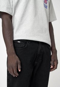 Camiseta gris de manga corta con logo rojo y azul, combinada con jeans de mezclilla negros. Enfócate en la textura de la tela y el ajuste relajado.