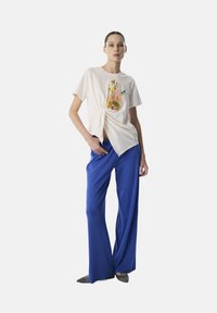 T-shirt en coton blanc avec un détail noué à la taille et un design de fruits colorés, associé à un pantalon bleu ample à côtes.