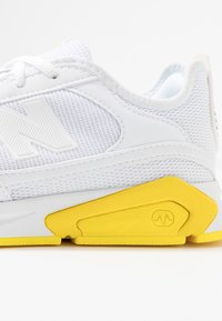 Chaussure de sport blanche avec tige en mesh, logo blanc proéminent et accent en caoutchouc jaune sur la semelle. Forme arrondie avec des détails texturés.