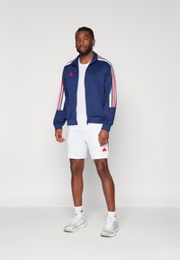 Giacca da tuta navy con strisce rosse e bianche, chiusura con zip, abbinata a pantaloni corti bianchi e scarpe da ginnastica grigie. Accenti del logo rosso su entrambi.