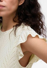 Helles gelbes Oberteil mit gestickten Eylet-Mustern, Rüschensäumen und einem hohen Ausschnitt. Strukturierter Stoff mit glatter Oberfläche.