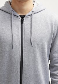 Šedá mikina s kapucí vyrobená z měkké tkaniny, s plným zipem, twill šňůrkami a kontrastním černým zipem.