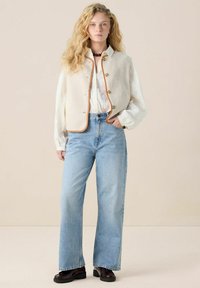 Gilet en polaire crème avec des bordures marron, chemise blanche, jeans larges bleu clair, et chaussures chunky bordeaux. Le mannequin se tient devant un fond neutre.