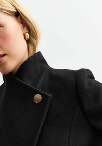 Cappotto di lana nero con colletto alto, caratterizzato da un bottone decorativo dorato. Texture liscia con vestibilità su misura e linee pulite.